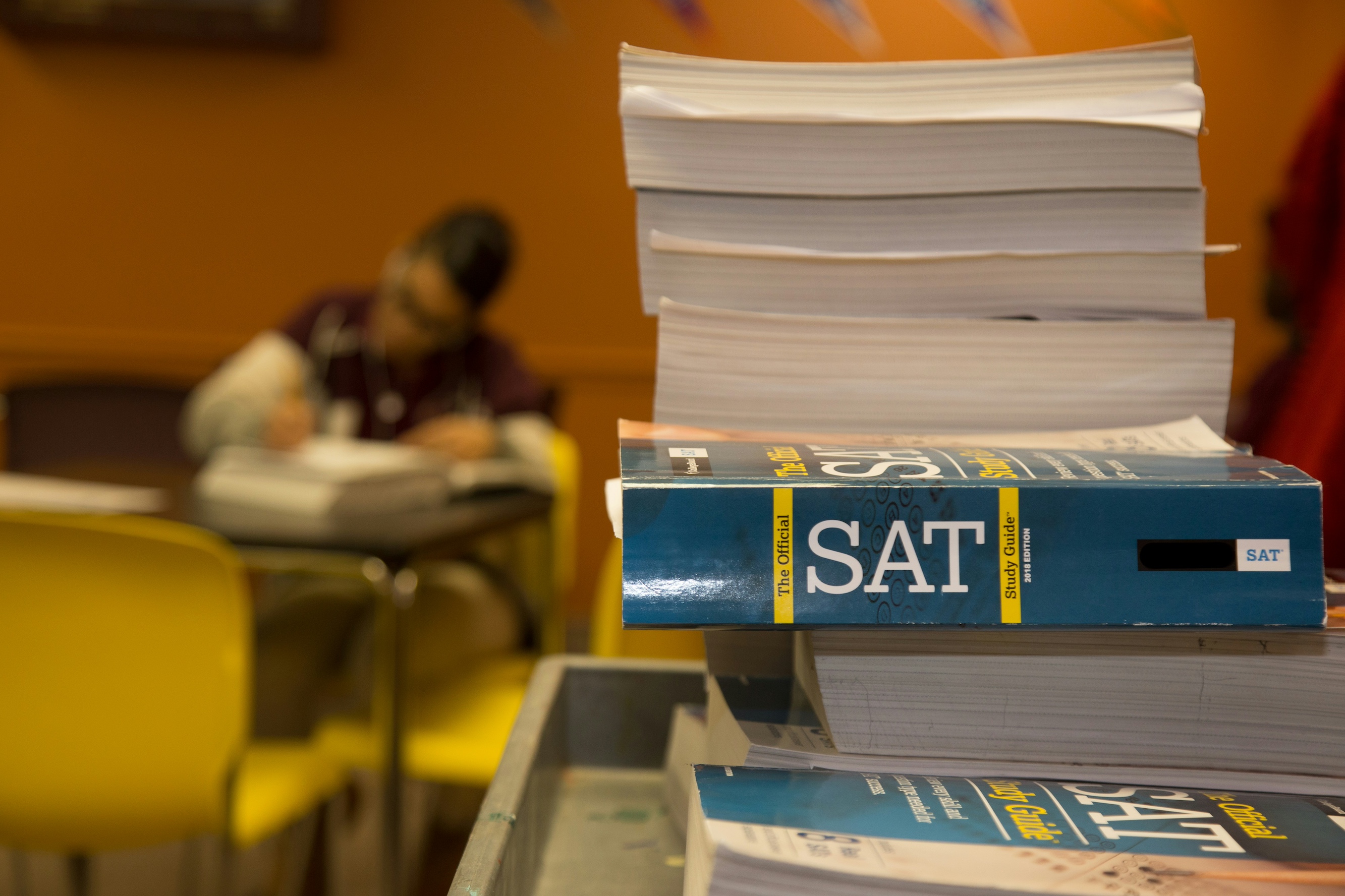 SAT test prep book stacked on a desk with student studying in the background.